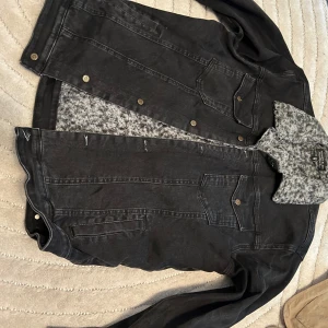Svart jeansjacka från Philipp Plein - Säljer en svart jeansjacka från Philipp Plein med grå teddyfoder och krage. Jackan har klassiska bröstfickor med knappar och coola detaljer. Perfekt för dig som vill ha en streetig look med extra edge.