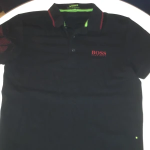 Svart Hugo Boss t-shirt med röd logga - Svart t-shirt från Hugo Boss med röd logga på bröstet och ryggen. Slim fit-modell med korta ärmar och klassisk krage med knappar. Materialet är bomull och har fuktavledande egenskaper. Snygga röda detaljer på kragen och ärmen ger extra stilpoäng.