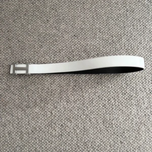 Vitt skärp med H-spänne - Snyggt vitt bälte med ett stort H-format metallspänne i silver. Bältet är slätt och stilrent, med svart insida och raka kanter. Perfekt accessoar för att lyfta din outfit med en clean och modern look.
