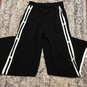 Svarta mjukisbyxor med vita revärer - Svarta mjukisbyxor med bred passform och vita dubbla revärer längs sidorna. Byxorna har resår i midjan och knytband längs benen som ger en cool detalj. Perfekta för en avslappnad och sportig stil.