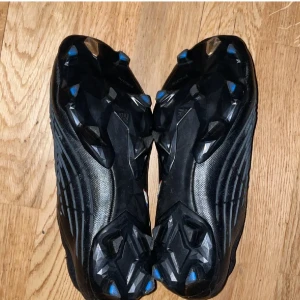 Adidas svarta fotbollsskor med ränder - Säljer ett par svarta Adidas fotbollsskor med klassiska tre ränder i vitt, blått och rött på sidan. Skorna har en strumpliknande ovandel och mönstrad sula med dobbar för bra grepp på planen. Tillverkade i syntetmaterial och designade för att sitta tajt på foten.