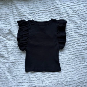 Svart topp med volangärmar Even&Odd - Supersnygg svart topp från Even&Odd med breda volangärmar som ger en cool statement-look. Kan rekommenderas till barn.