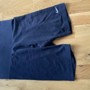 Svarta träningstights-shorts från Nike Storlek S - Svarta, tighta träningstights-shorts från Nike med hög midja och diskret Nike-logga på benet. Perfekta för gymmet eller löprundan, tillverkade i stretchigt syntetmaterial som andas och sitter skönt på kroppen.