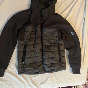 Svart shell höstjacka från C.P. Company - Säljer en svart pufferjacka från C.P. Company med ikonisk lins på ärmen. Jackan har quiltad front och insida, dragkedja och huva. Materialmix med polyester och syntet, perfekt för kyliga höstdagar. Snygg och modern look med normal passform. Passar både M och L.