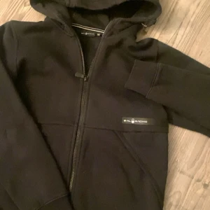 Svart Ziphoodie från Sail Racing - Svart hoodie från Sail Racing med dragkedja framtill och huva. Använd fåtall gånger