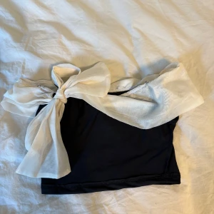 Svart topp med vit rosett - Supersnygg topp i svart med en stor vit rosett framtill. Off-shoulder design som ger en cool och trendig look. Materialet känns mjukt och bekvämt mot huden.