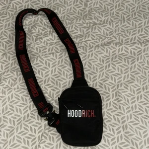 Svart axelväska från Hoodrich - Svart axelväska från Hoodrich med justerbart band där loggan står i rött. Väskan har dragkedja upptill och ett extra fack framtill. Snygg och praktisk för att bära det viktigaste, med cool urban vibe och tydlig branding.