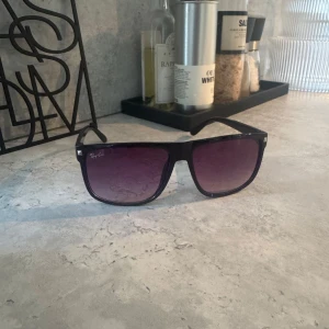Svarta Ray-Ban solglasögon - Snygga svarta Ray-Ban solglasögon med rektangulär form och mörka, tonade glas. Klassisk och stilren design med diskret logga på linsen. Perfekta för att lyfta din look med en cool vibe.