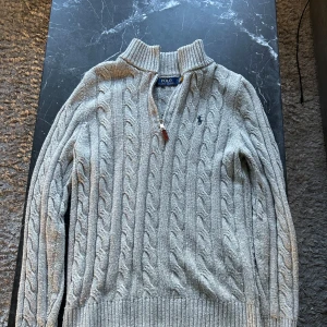 Half zip Ralph Lauren - Säljer en grå stickad tröja från Polo Ralph Lauren i storlek S. Tröjan har ett klassiskt kabelstickat mönster, ribbade muddar och krage samt en half zip-dragkedja framtill. Perfekt för dig som gillar stilren och tidlös design. Skick är 9/10. 