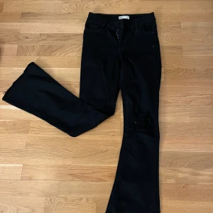 Svarta bootcut jeans med slitna knän från Gina Young  - Säljer ett par svarta jeans från Gina Young  med bootcut-modell och slitna detaljer vid knäna. Byxorna har klassisk femficksdesign och är tillverkade i ett stretchigt jeanstyg som sitter snyggt. Perfekta för dig som gillar en lite edgy stil.