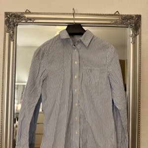 Blåvit randig skjorta från GANT - Snygg blåvit randig skjorta från GANT med klassisk krage och bröstficka med broderad logga. Skjortan har långa ärmar och knäppning framtill. 