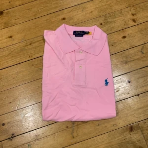 Rosa Polo Ralph Lauren t-shirt - Snygg rosa t-shirt från Polo Ralph Lauren med klassisk krage och två knappar framtill. Slim fit-modell med blå broderad logga på bröstet. Tillverkad i mjuk bomull som känns skön mot huden. Perfekt för dig som gillar stilrena och färgglada plagg. Storlek s.