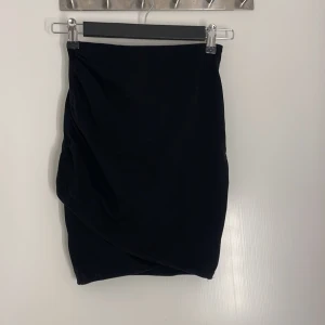 Svart omlottkjol i kort modell - En svart kortkjol med omlottdesign och lätt drapering på sidan. Kjolen har en stilren look och passar perfekt till sneakers eller boots. Enkel att matcha med olika toppar för en trendig outfit.