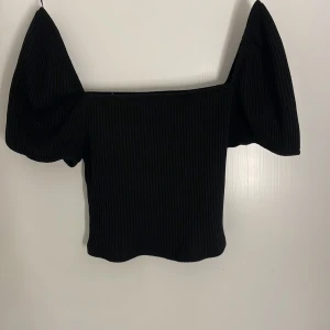 Svart ribbad topp med puffärm H&M - Svart ribbad topp från H&M med bred offshoulder-ringning och stora puffärmar. Toppen är croppad och har en tight passform som framhäver siluetten. Perfekt för dig som gillar trendiga detaljer och stilren look.