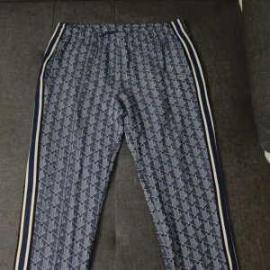 Blå mönstrade byxor från Lacoste - Snygga blå byxor från Lacoste med vitt och marinblått mönster över hela tyget. Byxorna har elastisk midja, vita och blå ränder längs sidorna och den klassiska krokodilloggan på höften. Perfekta för en sportig och trendig look.