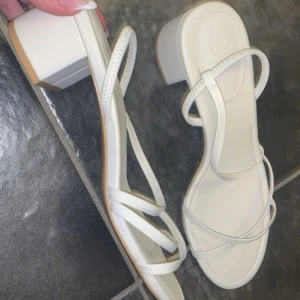 Vita sandaler med klack från H&M - Snygga vita sandaler från H&M med smala remmar och fyrkantig tå. Skorna har en låg blockklack och öppen tå, perfekta för sommarens alla outfits. Tillverkade i syntetmaterial med stilren och minimalistisk design.