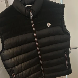 Svart dunväst från Moncler - Snygg svart dunväst från Moncler med hög krage och klassisk logga på bröstet. Västen har dragkedja framtill med Monclers signaturband längs sidorna och två fickor med dragkedja. Perfekt för lager-på-lager och streetstyle.
