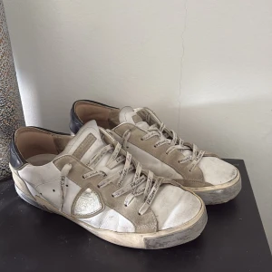 Philippe Model vita sneakers med svarta detaljer - Snygga sneakers från Philippe Model i vitt skinn med beige mockadetaljer och svarta hälkappor. Sulan är ganska slitna och har ett hål på höger sko, går att laga för bra pris! Skorna har rund tå, platt sula och snörning med logotyp på skosnörena. Klassisk streetstil med coola kontraster och logga bak på hälen.