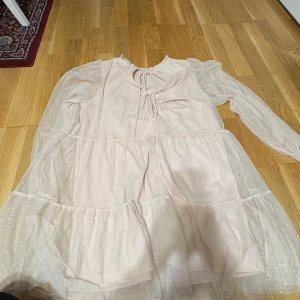 Ljusrosa volangklänning med tyll - Supersöt ljusrosa kortklänning med volanger och transparenta långa ärmar i tyll. Klänningen har små prickar i tyllen, knytning vid halsen och en luftig, flowy passform. Perfekt för dig som gillar romantisk och feminin stil.