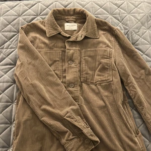 Beige manchesterjacka These Glory Days - Säljer en beige overshirt i manchester från These Glory Days. Jackan har klassisk krage, knäppning framtill, bröstficka och långa ärmar. Perfekt för lager-på-lager och ger en chill vibe till din outfit.