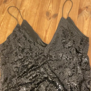 Svart paljettlinne med smala axelband - Snygg svart linne med smala axelband och glittriga paljetter över hela ytan. Linnet har en v-ringad hals och är kort i modellen, perfekt för dig som vill sticka ut med lite extra bling. Materialet känns mjukt och har ett satintyg på insidan för extra komfort.