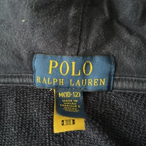 Marinblå hoodie från Polo Ralph Lauren - Snygg marinblå hoodie från Polo Ralph Lauren med vit logga på huvan och klassisk broderad polospelare på bröstet. Tröjan har dragkedja framtill, huva och är tillverkad i mjuk bomull. Perfekt för en chill och sportig look.