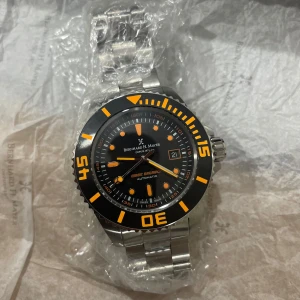 Bernhard H. Mayer Wave Breaker klocka - Säljer en Bernhard H. Mayer Wave Breaker automatisk klocka med svart urtavla och orange detaljer. Klockan har robust silverfärgad länk i metall och levereras i originalbox med extra svart gummiband och verktyg. Perfekt för dig som gillar sportig och stilren design.Helt ny klocka 