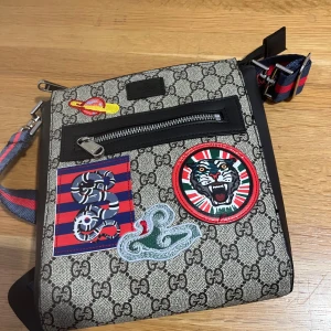 Gucci axelväska med patchar och GG-mönster - Gucci axelväska i beige med klassiskt GG-mönster, dekorerad med färgglada patchar som tiger, orm och planet. Väskan har svart skinnkant, dragkedja framtill och justerbar axelrem i blått och rött. Perfekt för dig som vill sticka ut med en unik designeraccessoar.