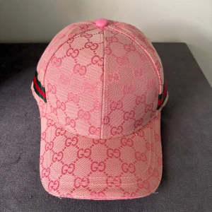 Rosa Gucci keps med monogram - Säljer en snygg rosa keps från Gucci med klassiskt GG-monogram över hela kepsen. Kepsen har justerbar rem bak i rosa läder och gröna/röda ränder på sidan. Materialet är bomull och detaljer i läder. Perfekt för dig som vill sticka ut med lyxig streetstyle.