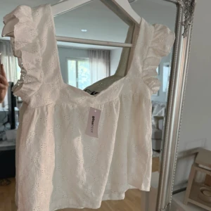 Vit blus med volang från Gina Tricot - Supersöt vit blus från Gina Tricot med breda axelband och volangdetaljer. Blusen har broderade blommor i tyget och en luftig, lätt passform. Ny med lapp