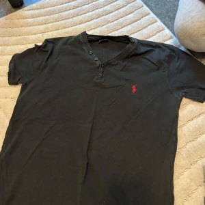 Svart t-shirt från Ralph Lauren - Svart t-shirt från Ralph Lauren med korta ärmar och v-ringning med tre knappar. Klassisk röd Polo-logga broderad på bröstet. Skön bomullskvalitet och normal passform, perfekt för chill dagar. Snygg och enkel stil som alltid funkar.