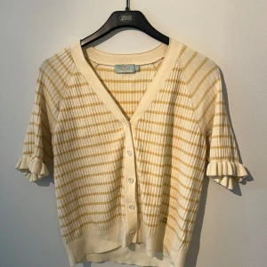 Randig kofta från Morris Lady - Superfin kofta från Morris Lady i beige och ljusgul med ribbad struktur och korta ärmar med volangdetalj. V-ringad med knappar framtill och mjukt stickat material. Perfekt för dig som gillar en chill och söt stil.