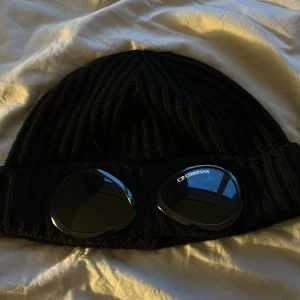 Svart mössa från C.P. Company - Svart mössa från C.P. Company med fina linser framtill. Mössan är tillverkad i mjukt material och har en uppvikt kant. 
