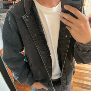 Svart jeansbomberjacka från Levi's - Säljer en mörkgrå bomberjacka från Levi's i jeansmaterial. Jackan har ribbade muddar, tryckknappar framtill och två fickor på sidorna. Oversized passform och klassisk bomberkrage ger en chill vibe. Perfekt för dig som gillar streetwear och vill ha en stilren look. Jackan är aldrig använd och har fortfarande prislapp på. Nypris 1020 kr 