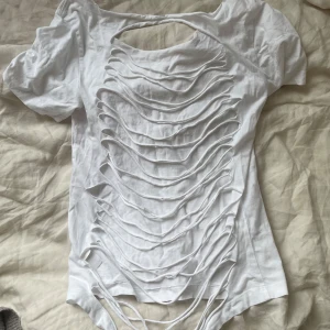 Vit t-shirt med slitsad design - Säljer en vit t-shirt med korta ärmar och unik slitsad design på baksidan. T-shirten har flera horisontella öppningar som ger en edgy och cool look. Materialet känns som bomull och passformen är normal. Perfekt för dig som vill sticka ut med din stil.