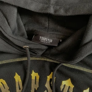 Svart hoodie från Trapstar med tryck - Svart hoodie från Trapstar med huva och snörning. Framsidan har ett stort tryck i guldigt/gult med gotisk stil. Hoodien har synliga sömmar i kontrasterande färg och känns street och edgy. Perfekt för dig som gillar statement-plagg.