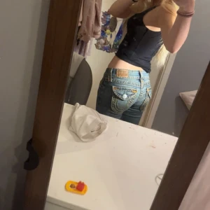 True Religion jeans med kontrastsömmar - Snygga blå jeans från True Religion med ikoniska kontrastsömmar i gult och detaljerade bakfickor med stora knappar. Klassisk femficksmodell i denim med raka ben och låg midja. Perfekta för dig som vill sticka ut med en streetig vibe. Har dock en liten skavank vid ena fickan men inget som syns se bilderna. 