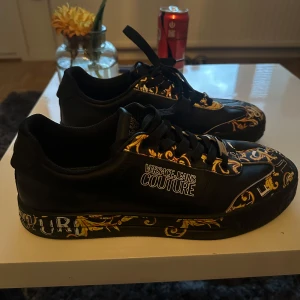 Versace Jeans Couture sneakers svart/guld - Riktig feta sneakers från Versace Jeans Couture i svart med guldiga och vita barockinspirerade detaljer och logotyptryck. Skorna har snörning, platt sula och är tillverkade i skinnliknande material. Perfekta för dig som vill sticka ut med en lyxig och trendig look använda bara några gånger 