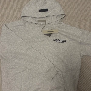 Grå Essentials Fear of God hoodie - Snygg ljusgrå hoodie från Fear of God Essentials med svart logga på bröstet och stor text på ryggen. Oversized passform, huva och ficka framtill. 