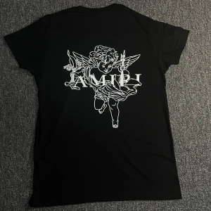 Svart AMIRI t-shirt med ängeltryck - Svart t-shirt från AMIRI med stort vitt ängeltryck och logga på ryggen samt färgstänk och AMIRI-tryck framtill. Klassisk passform med korta ärmar och rund hals. Tillverkad i mjuk bomull, perfekt för en cool streetwear-look.