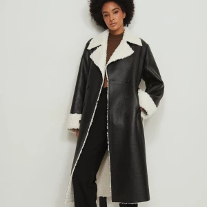 Svart lång pälsjacka med vita detaljer - Lång svart pälsjacka med vita teddy-detaljer på krage, ärmslut och insida. Dubbelknäppt framtill, bred krage och markerade sömmar. Jackan har en cool oversized vibe och är perfekt för kalla dagar. Materialet är skinnimitation med mjuk teddy på insidan.