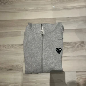 Grå hoodie Comme des Garçons Play - Grå hoodie med dragkedja från Comme des Garçons Play. Ikoniskt svart hjärta med ögon på bröstet och stort hjärta på ryggen. Mjuk bomull, huva och långa ärmar. Perfekt för dig som gillar streetstyle och vill sticka ut med en klassiker.