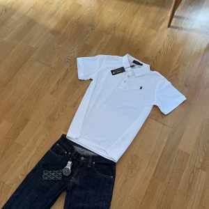 Vit pikétröja från Polo Ralph Lauren - Klassisk vit pikétröja från Polo Ralph Lauren med korta ärmar och broderad logga på bröstet. Tröjan har krage, knappar framtill och är tillverkad i bomull för en fräsch och stilren look. Perfekt till jeans eller shorts.