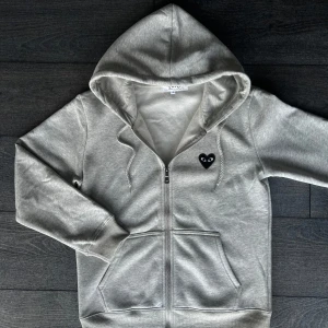 Grå CDG hoodie - Grå hoodie med dragkedja från Comme des Garçons Play. Ikoniskt svart hjärta med ögon på bröstet och stort tryck på ryggen. Mjuk bomull, huva med snörning och två fickor framtill. Perfekt för en chill och stilren look.