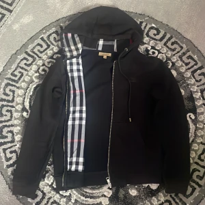 Burberry hoodie - Svart hoodie från Burberry med klassiskt rutigt foder i huvan och längs insidan. Tröjan har huva med snörning, lång ärm och dragkedja framtill. Perfekt för dig som gillar stilrena designer med en touch av lyx.