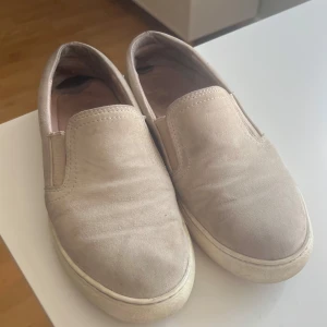 Beige/rosa sneakers i mocka - Snygga slip-on sneakers i beige mocka med vit platt sula. Skorna har rund tå och elastiska paneler på sidorna för enkel påtagning. Perfekt för dig som gillar enkel och clean stil. Passar till jeans, shorts eller kjol för en chill look.