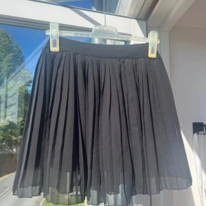 Svart plisserad kjol från NA-KD - Supersnygg svart plisserad kortkjol från NA-KD i storlek S. Kjolen har ett brett elastiskt midjeband och är gjord i ett lätt, flowy material som ger en cool och trendig vibe. Perfekt att matcha med en crop top eller hoodie för en chill look.