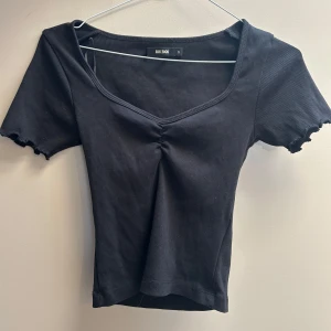Svart ribbad topp från Bik Bok - Svart ribbad topp från Bik Bok med v-ringning och korta ärmar. Toppen har en snygg rynkdetalj framtill och vågiga kanter vid ärmslut. Perfekt för en trendig och avslappnad stil.