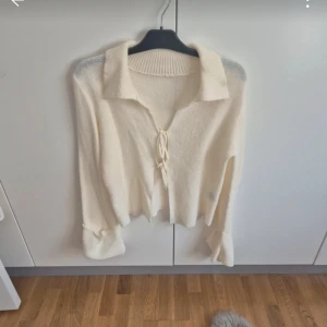 Beige stickad kofta med snörning - Superfin beige stickad kofta med krage och snörning framtill. Tröjan har långa ärmar med bred uppvikning och en croppad passform som ger en trendig look. Perfekt att styla med jeans eller kjol för en chill vibe.