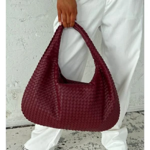 Vinröd väska Designbysi must have bag  - Helt ny! Säljer en stor och mjuk axelväska i vinrött skinn med flätad design. Väskan har en rundad, slouchy form och bred axelrem som gör den enkel att bära. Perfekt för dig som gillar att ha med dig mycket och vill sticka ut med en snygg accessoar.  Får plats med en 13 tum dator 